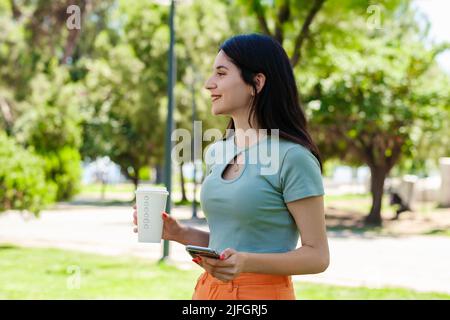 Brunette Millennial Frau trägt türkisfarbenes T-Shirt im Stadtpark, im Freien hält Kaffee zum Mitnehmen Tasse und mit Handy, während weg schauen. Stockfoto