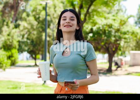 Porträt einer fröhlichen Frau, die türkisfarbenes T-Shirt im Stadtpark trägt, im Freien mit Kaffeebecher zum Mitnehmen hält und ein Mobiltelefon verwendet, während sie den ansieht Stockfoto