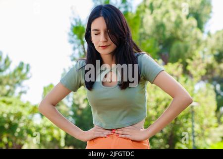 Brunette tausendjährige Frau trägt türkisfarbenen Tee im Stadtpark, im Freien mit der Hand auf dem Bauch, weil Verdauungsstörungen, schmerzhafte Krankheit Gefühl unwohl. Schmerzen Stockfoto