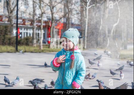 Lachendes Mädchen in einem Frühlingspark. Ein Kind mit langen Haaren, die in Zöpfe geflochten sind. Ein Mädchen in einer blauen Jacke. Eine Taubenschar ist im Hintergrund. Childh Stockfoto