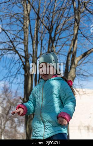 Ein lachendes Mädchen posiert für die Kamera im Park. Ein Kind mit langen Haaren, die in Zöpfe geflochten sind. Mädchen in einer blauen Jacke in einem Frühlingspark. Kindheit. Tre Stockfoto