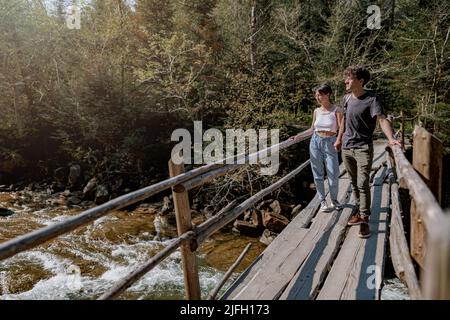 Ein paar Wanderer überqueren den Fluss durch eine Holzbrücke in den Bergen. Schnelles Wasser. Trekking. Stockfoto