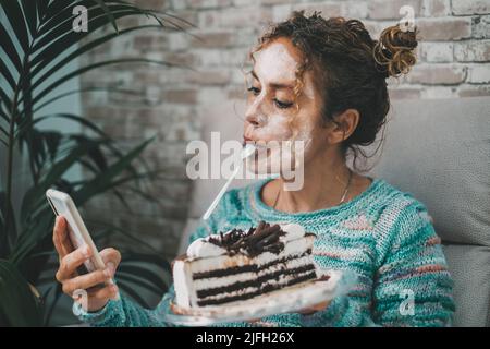 Junge, reife Frau, die zu Hause ein Mobiltelefon benutzt und einen Eiskuchen isst. Weiße Schönheitscreme-Behandlung im Gesicht. Indoor Freizeit weibliche Leute Aktivität.. Stockfoto
