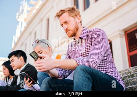 Rotschopf Kerl mit Handy. Freudiger, unrasierter Mann, der auf der Treppe mit dem Mobiltelefon telefoniert Stockfoto