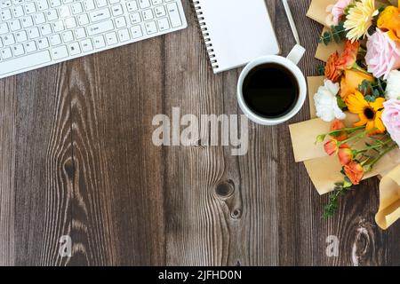 Schreibtisch mit Computertastatur, gemischtem Blumenstrauß und einer Tasse Kaffee auf Holzhintergrund Stockfoto