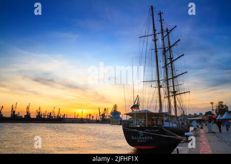 Blick auf den Hafen und die Uferpromenade von Varna bei Sonnenuntergang, Segelschiff mit Hafenkranen im Hintergrund, an der Schwarzmeerküste Bulgariens Stockfoto