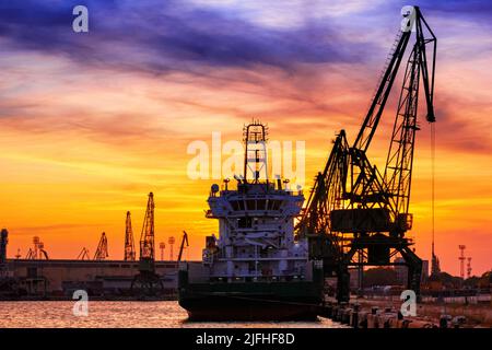 Silhouetten von Hafenkranen und Frachtschiffen im Hafen von Varna bei Sonnenuntergang, an der bulgarischen Schwarzmeerküste Stockfoto