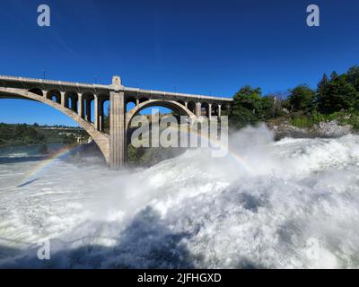 Sintflutartige Strömung über den Lower Spokane Falls im Stadtzentrum von Spokane nach heftigen Regenfällen am hellen, sonnigen Sommermorgen mit Regenbogen und der Monroe Street Bridge. Stockfoto