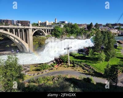 Sintflutartige Strömung über den Lower Spokane Falls im Stadtzentrum von Spokane nach heftigen Regenfällen am hellen, sonnigen Sommermorgen mit Regenbogen und der Monroe Street Bridge. Stockfoto