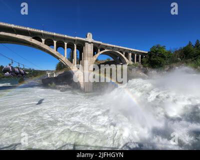 Sintflutartige Strömung über den Lower Spokane Falls im Stadtzentrum von Spokane nach heftigen Regenfällen am hellen, sonnigen Sommermorgen mit Regenbogen und der Monroe Street Bridge. Stockfoto