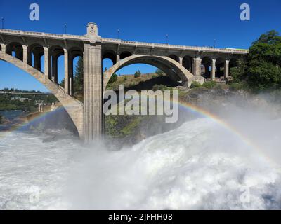 Sintflutartige Strömung über den Lower Spokane Falls im Stadtzentrum von Spokane nach heftigen Regenfällen am hellen, sonnigen Sommermorgen mit Regenbogen und der Monroe Street Bridge. Stockfoto