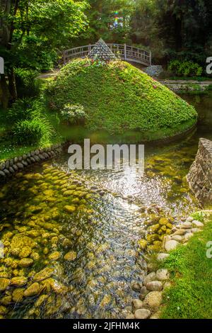 Der Teich mit Steinbänken und einer Art Azaleen Büschen Pyramide und japanische Brücke dahinter und Sonne glänzte in Wasserspiegelungen in Albert Kahn Stockfoto