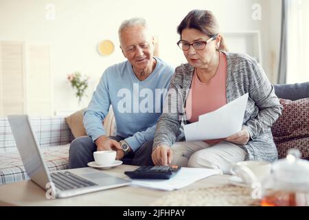 Ältere Ehepaare in legerer Kleidung sitzen auf dem Sofa im Wohnzimmer und sehen sich die Papiere an, während sie gemeinsam die monatlichen Ausgaben berechnen Stockfoto