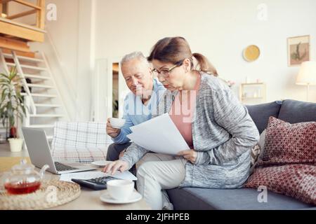 Seriöse ältere Frau mit Brille sitzt auf dem Sofa und schaut sich Papiere an, während sie zusammen mit ihrem Mann die Haushaltsfinanzen analysiert Stockfoto