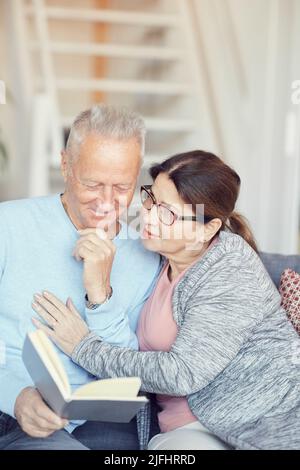 Lächelnder grauhaariger Mann in blauem Pullover, der das Kinn berührt, während er mit seiner älteren Frau zu Hause ein Buch liest Stockfoto
