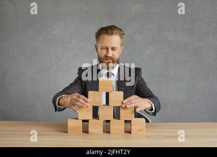 Der Geschäftsleiter sitzt am Schreibtisch und baut eine Pyramide aus Holzblöcken Stockfoto