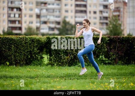 Junge Frau in Tanktop und Leggings joggen im öffentlichen Park neben einem Wohngebiet mit Hochhäusern. Stockfoto