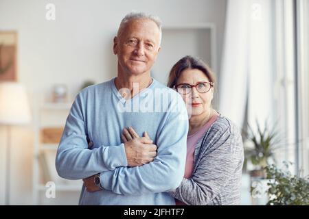 Lächelndes, wunderschönes Ehepaar, das im Wohnzimmer steht und sich umarmt, während es für ein Porträt posiert Stockfoto