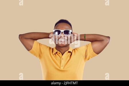 Cooler, glücklicher afroamerikanischer Mann in einer Sonnenbrille, der die Hände hinter dem Kopf auf hellbeigem Hintergrund hält. Stockfoto