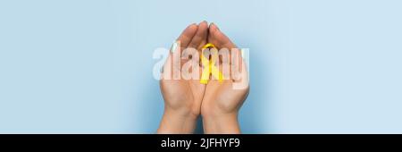 Gelbes Goldband in den Händen eines Mädchens auf blauem Hintergrund. Kinderkrebskonzept. Monat der Sensibilisierung. Kinderkrebstag Februar 15. Internet Stockfoto