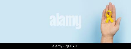Gelbes Goldband in der Hand einer Frau auf blauem Hintergrund. Kinderkrebskonzept. Monat der Sensibilisierung. Kinderkrebstag Februar 15. Internet Stockfoto