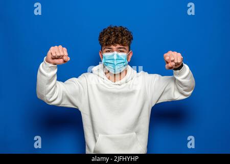 Das Porträt eines gutaussehenden Mannes in einer medizinischen Schutzmaske, der mit erhobenen Armen steht, zeigt starke Bizeps. Isolierter Hintergrund. Das Konzept des Aufenthalts Stockfoto