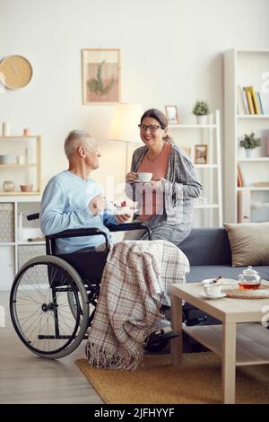 Fröhliche ältere Frau in einer Brille, die Tee trinkt und lacht, während sie dem Ehemann, der im Rollstuhl sitzt und Kuchen isst, eine lustige Geschichte erzählt Stockfoto