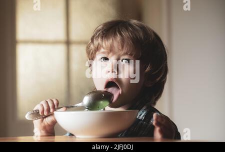 Kleines Kind sitzt auf dem Tisch essen mit lustigen Ausdruck auf dem Gesicht. Leckeres Kinderfrühstück. Leckeres Kinderfrühstück. Fröhliches Baby isst selbst Nahrung Stockfoto