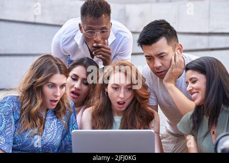 Eine Gruppe multiethnischer Menschen blickt auf den Laptop-Bildschirm mit einem schockierten Gesichtsausdruck, während sie draußen auf einer Treppe sitzen. Urbaner Lebensstil, Technologie und Freundschaftskonzept. Stockfoto
