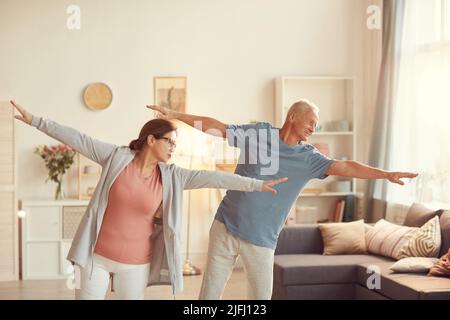 Inhalt Seniorenpaar im aktiven Ohr steht im Wohnzimmer und streckt die Arme aus, während sie zu Hause gemeinsam trainieren Stockfoto