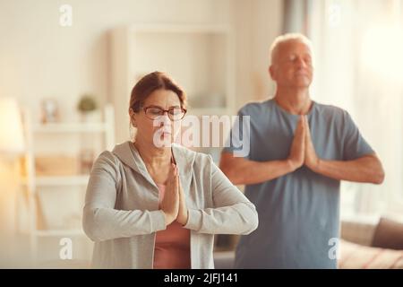Ruhiges, gealtertes Paar konzentrierte sich auf Meditation, die im Wohnzimmer stand und Namaste-Geste machte Stockfoto