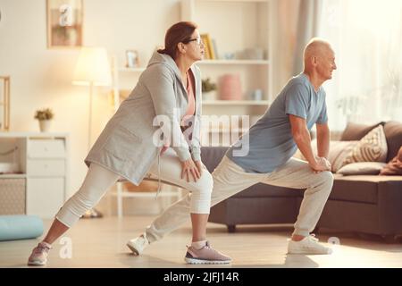 Energisches Senior-Paar in Sportswear, das sich beim Training zu Hause auf die Knie stützt und dabei die Beine streckt Stockfoto