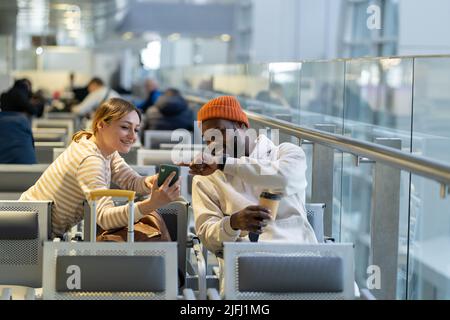 Ein glückliches, junges internationales Paar, das in der Lounge des Flughafens sitzt und sich mit einander unterhält und auf die Abreise wartet. Stockfoto