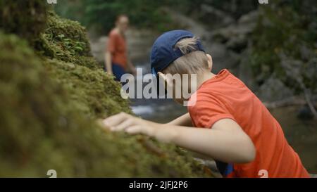 Konzept des Reisens. Kreativ. Rückansicht eines Jungen in einem Sommerwald, der vorsichtig am Flusslauf entlang läuft und moosigen Berghang berührt. Stockfoto