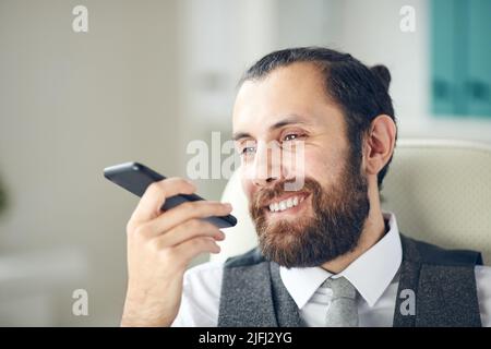 Positiv schöner bärtiger Geschäftsmann, der auf einem Bürostuhl sitzt und Smartphone benutzt, während er eine lustige Botschaft für einen Kollegen aufzeichnet Stockfoto