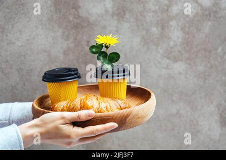 Zwei gelbe Pappbecher für Kaffee oder Tee und frische Croissants auf einem Holztablett. Bestellen Sie alles in den Händen der Frauen. Im Deckel eines Behälters befindet sich eine Blume aus Delelion für ein romantisches Frühstück. Platz für Text kopieren Stockfoto