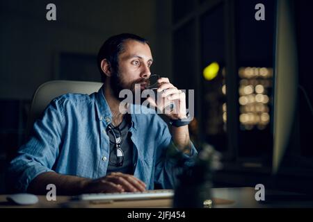 Seriös nachdenklicher junger bärtiger Mann in legerem Hemd, der im Nachtbüro sitzt und Wasser trinkt, während er den Computer benutzt Stockfoto