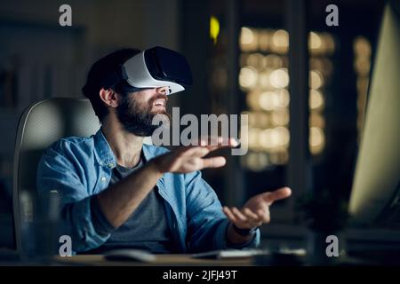 Aufgeregt junger bärtiger Mann, der vor dem Computer sitzt und die Hände während des Testens der neuen App über ein VR-Gerät gestikelt Stockfoto