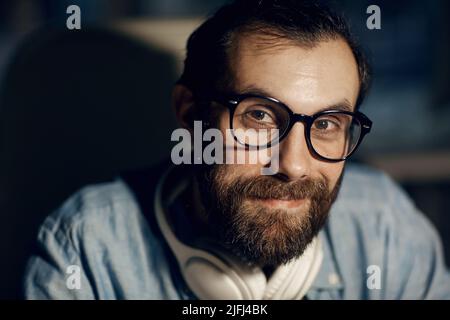 Portrait of content Hipster junger Mann mit Schnurrbart und Bart tragen Brillen sitzen in dunklen Raum Stockfoto