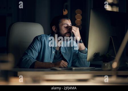 Müde junge Hipster Computer Programmierer in Casual Shirt sitzen am Tisch und reiben Brücke der Nase während der Arbeit spät im Büro Stockfoto