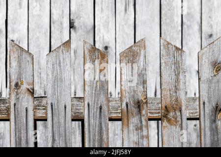 Holzzaun aus spitzen Brettern mit blasser hölzerner Gebäudewand in verschwommenem Hintergrund. Nahaufnahme verwitterter Schutzzäune mit Lücken zwischen brauner Planke. Stockfoto