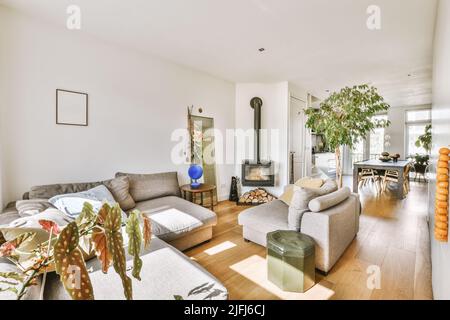 Großes Ecksofa mit weichen Kissen, in der Nähe des Kamins in einem hellen Wohnzimmer mit Topfpflanzen, neben dem Esszimmer und der offenen Küche Stockfoto
