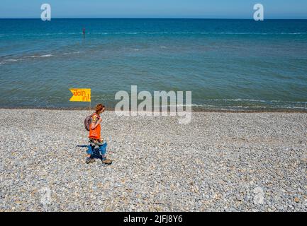 Frau, die mit einer gelben Mitfahrflagge am Kiesstrand entlang geht, in Sheringham, Norfolk, England. Stockfoto