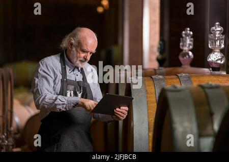 Ein im Kaukasus erfahrener Winzer überprüft eine Weinalterung in Fässern mit einem Touchpad-Tablet. Traditionelles und modernes Technologiekonzept. Stockfoto