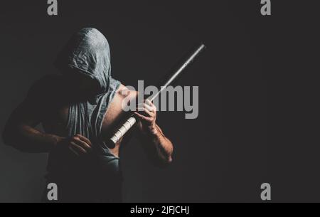 Gefährlicher Mann in der Maske mit Baseballschläger bereit zum Kampf. Ein aggressiver muskulöser Kapuzenmann hält einen Baseballschläger in der Hand Stockfoto