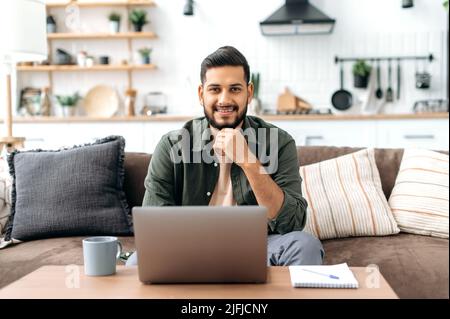 Gutaussehender selbstbewusster attraktiver, positiver indischer oder arabischer junger Mann, in lässiger, stilvoller Kleidung, sitzt zu Hause im Wohnzimmer auf dem Sofa, mit Laptop, schaut freundlich lächelnd auf die Kamera Stockfoto