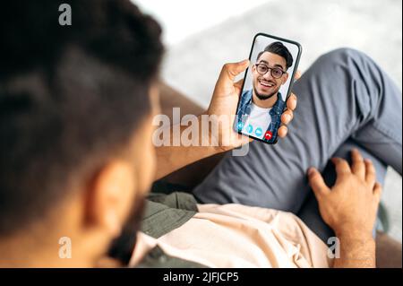 Blick auf ein Telefon mit indischen oder arabischen Kerl. Zwei Männer sprechen auf einer Online-Videokonferenz mit einer Anwendung und einem Smartphone, sind in einem Abstand voneinander, lächelnd freundlich Stockfoto