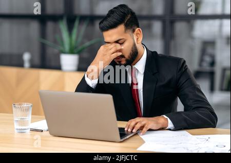 Müde erschöpfter indischer oder arabischer Geschäftsmann in einem Anzug, sitzen am Arbeitsplatz im Büro, reiben seine Nasenbrücke, Gefühle Müdigkeit nach langer Zeit Arbeit mit Laptop, überarbeitet, brauchen Ruhe, Kaffeepause Stockfoto