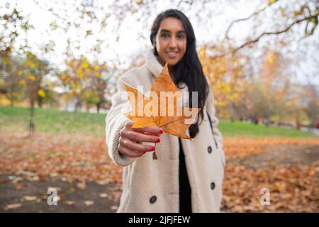 Mädchen hält ein orangefarbenes Blatt, das gerade vom Baum gefallen ist, in einem öffentlichen Park, konzentrieren Sie sich auf den Urlaub, Herbstkonzept Stockfoto