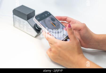 Weibliche Hand hält Smartphone für ein Foto mit dem RI eines Refraktometers, um die Art von Schmuck zu bestätigen. Stockfoto